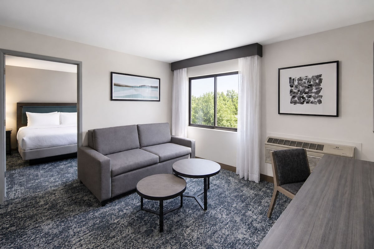 A hotel room with a bed, a grey sofa, a small round table, and two framed pictures on the walls.