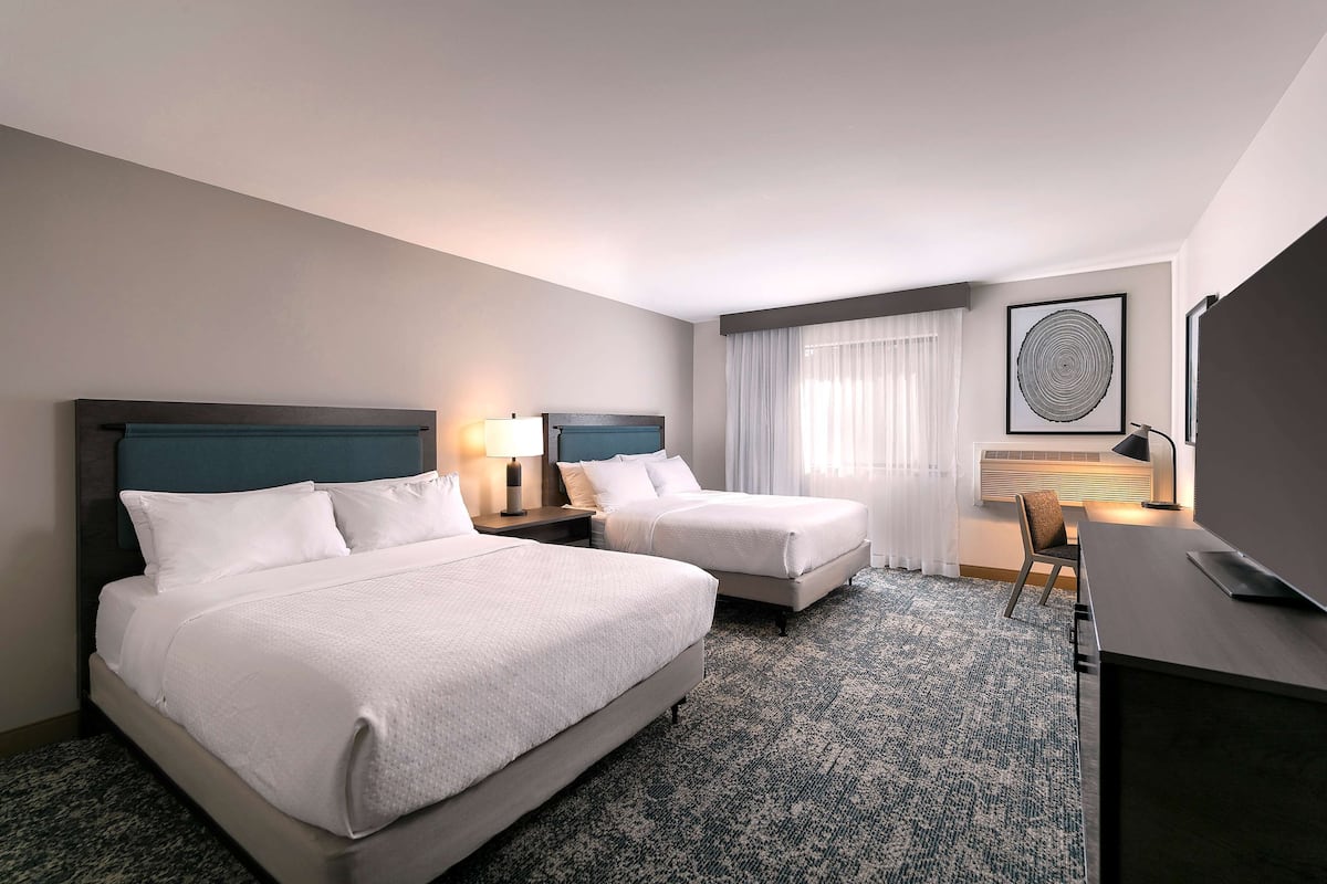 A hotel room with two beds, a desk, a chair, a lamp, a framed picture, and a window with curtains.