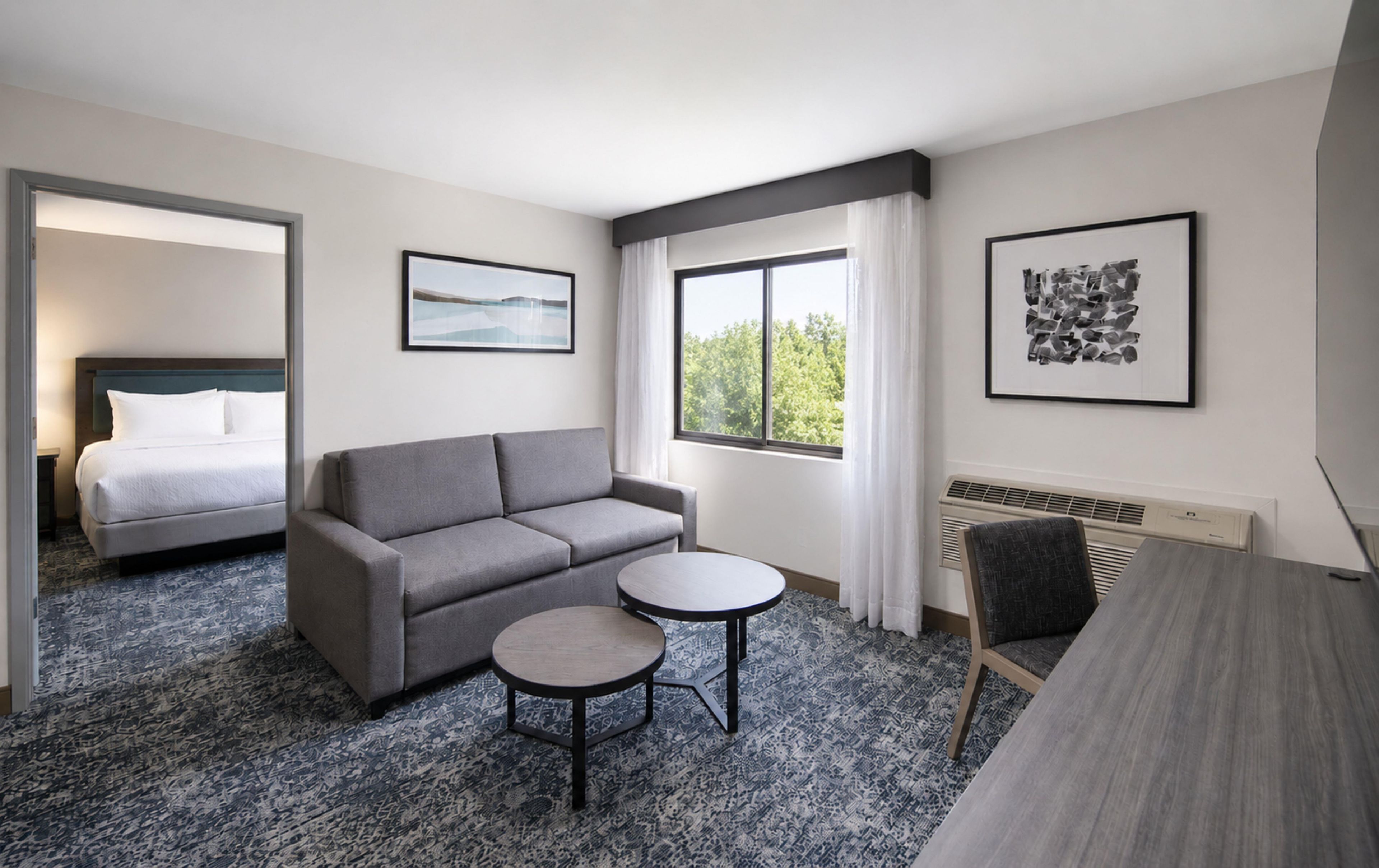 Une chambre d’hôtel avec un lit, un canapé gris, une petite table ronde et deux fenêtres.