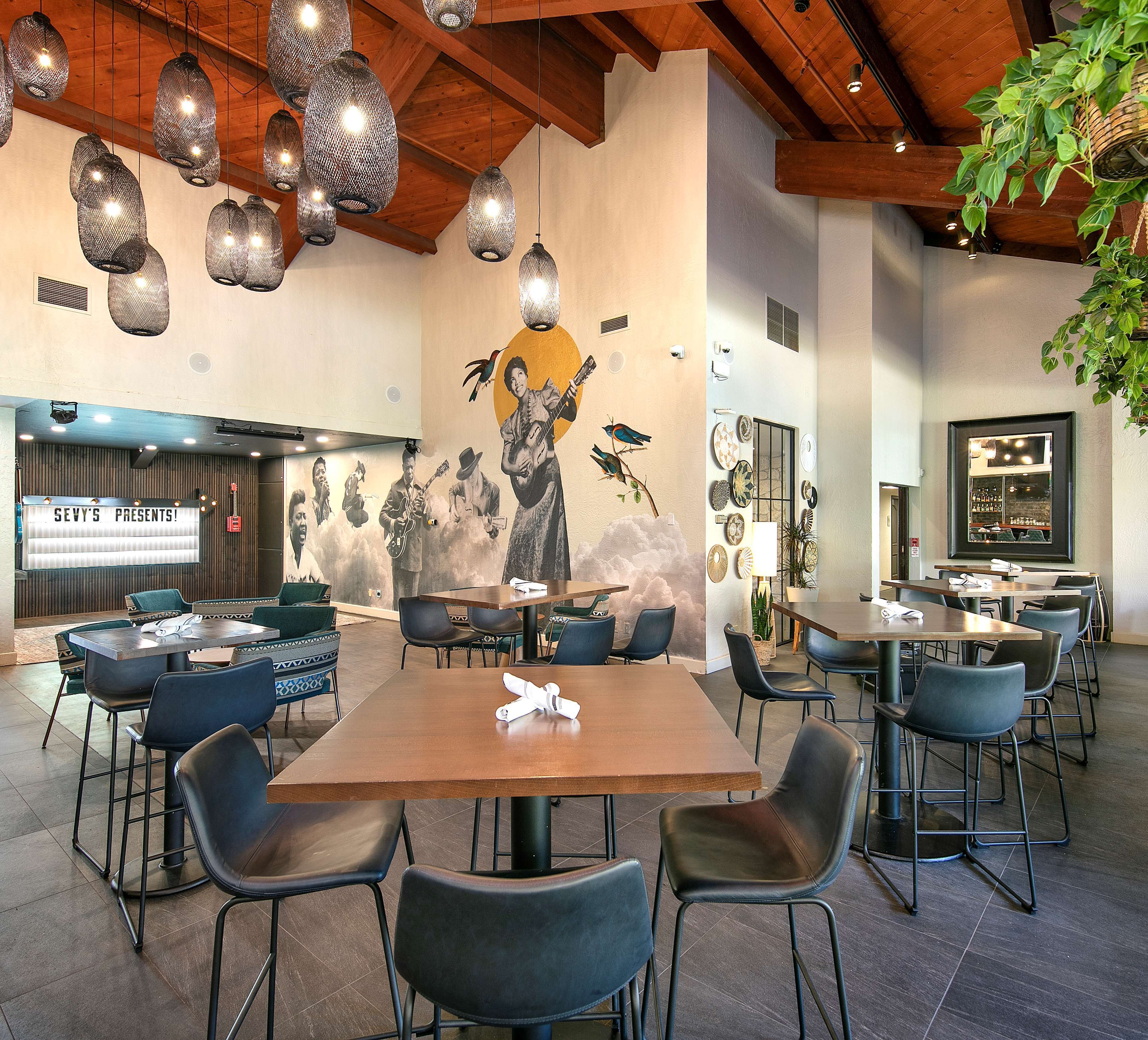 A modern restaurant with wooden beams, hanging pendant lights, and a mural of musicians. Tables and chairs are arranged for dining.