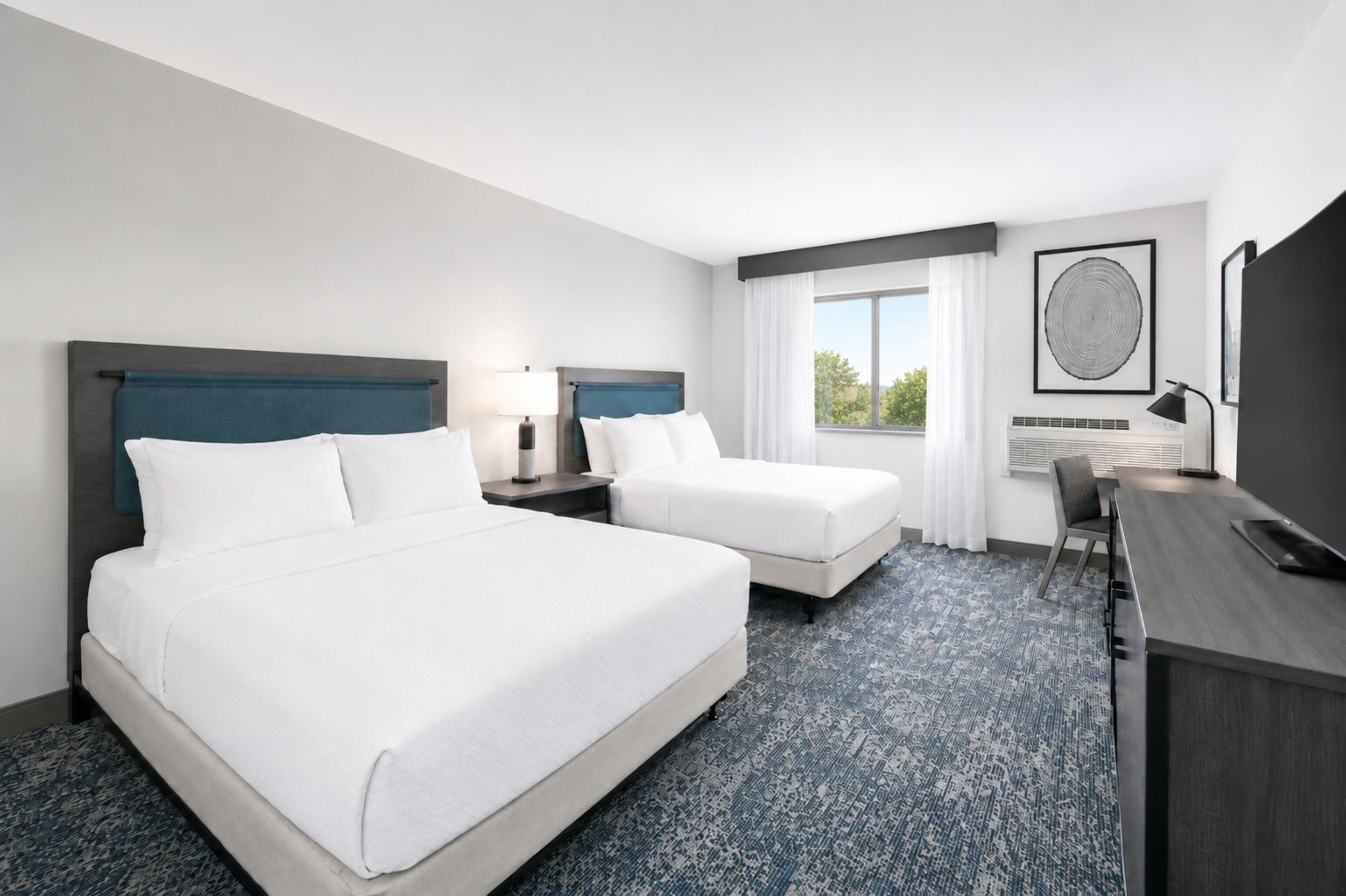 Quarto de hotel com duas camas, uma escrivaninha, uma cadeira e uma janela grande com cortinas.