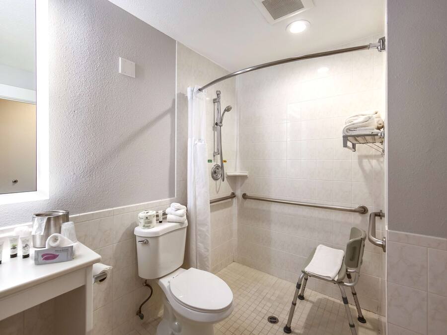 Un baño con inodoro, lavamanos con artículos de aseo, ducha con barra de apoyo y banco accesible para sillas de ruedas.