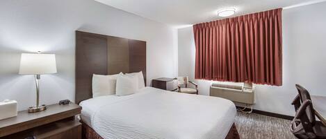 Habitación de hotel con una cama grande, un escritorio con lámpara, una silla, una mesita y una ventana con cortinas.