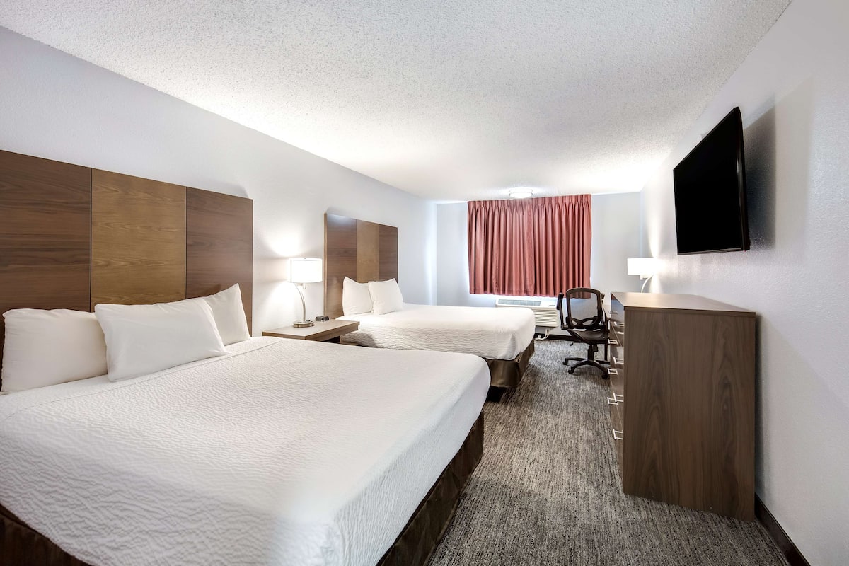 A hotel room with two beds, a desk, a chair, a flat-screen TV, and a window with curtains.