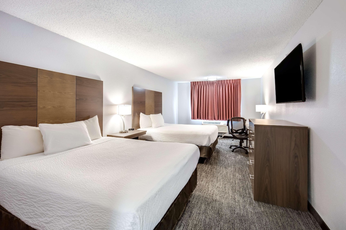 A hotel room with two beds, a desk, a chair, a flat-screen TV, and a window with curtains.