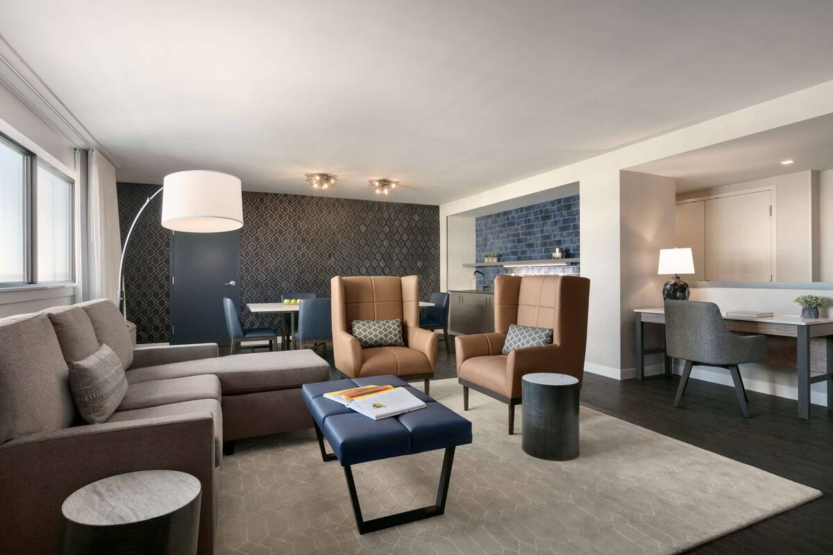 A modern living room with a sofa, armchairs, a coffee table, and a dining area with a kitchen in the background.