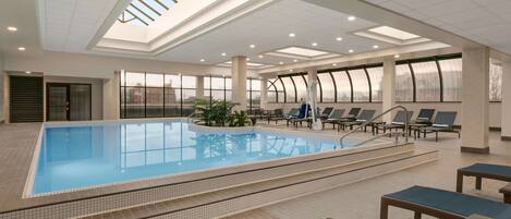 A large indoor swimming pool with clear glass walls, sunlit ceiling, and lounge chairs around the pool.
