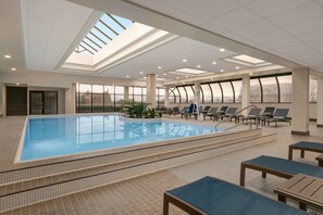Una amplia piscina interior con paredes de vidrio transparente, techo iluminado por el sol y sillones de descanso alrededor.