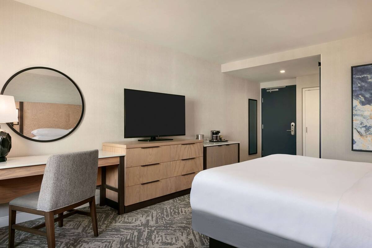 A hotel room with a bed, a desk with a chair, a television, a dresser, and a door.