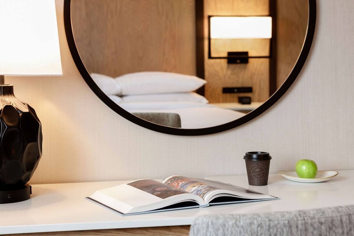 A hotel room with a bed, a bedside lamp, a coffee cup, a book, and a green apple on a bedside table.
