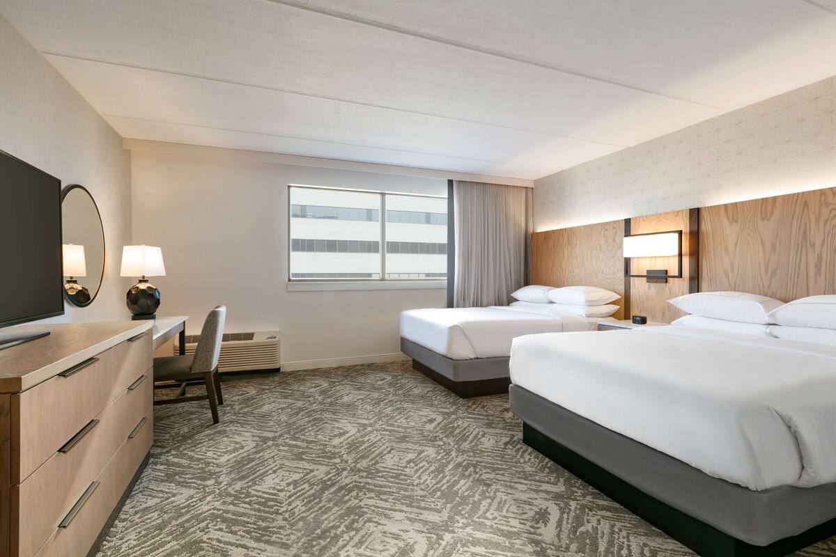 A hotel room with two beds, a desk with a chair, a TV, and a large window with curtains.