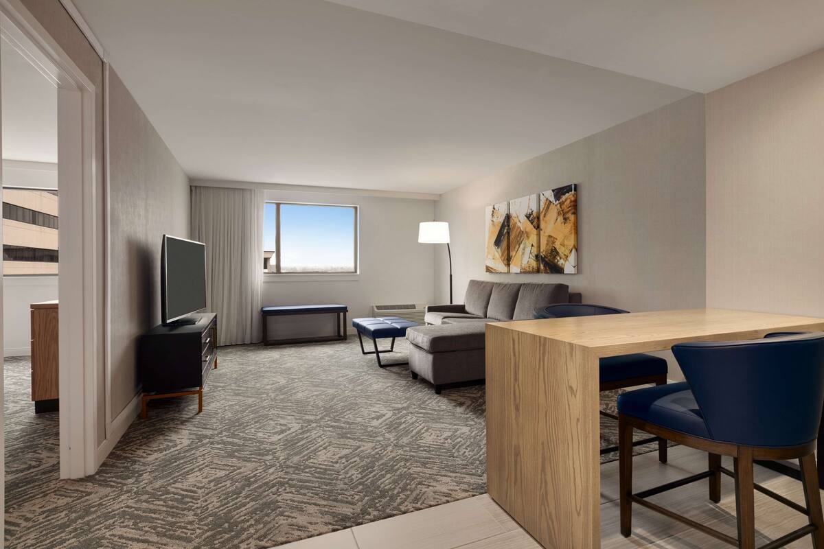 A modern hotel room with a large window, a flat-screen TV, a sofa, a dining table, and a lamp.