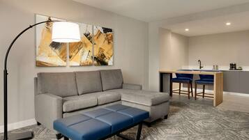A modern living room with a gray sectional sofa, a blue bench, and a floor lamp. There is a kitchen area with a bar counter and two blue bar stools.