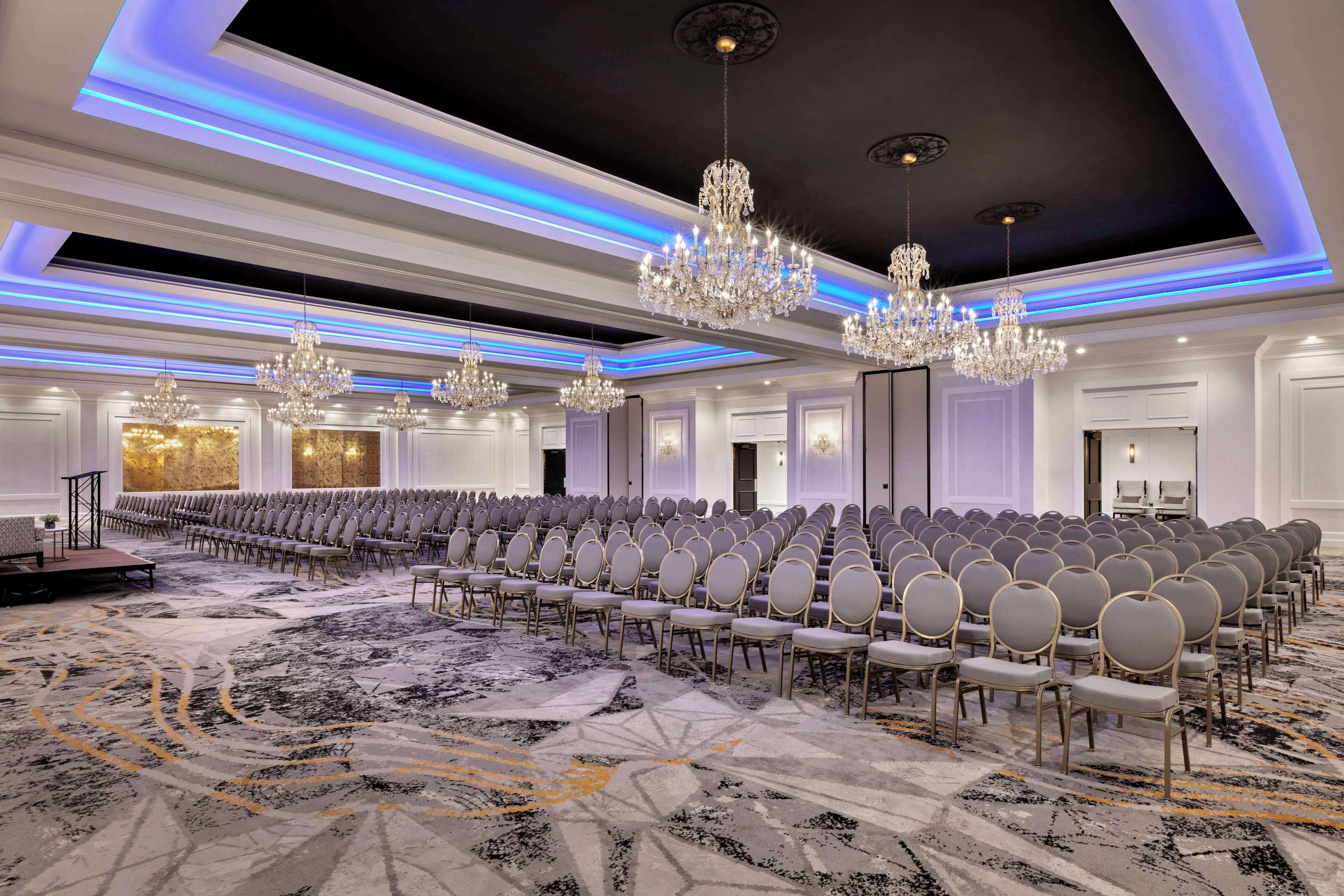 A large conference room with rows of chairs, chandeliers, and a stage.