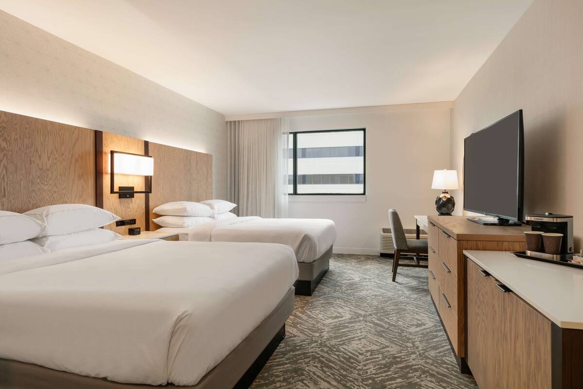 A hotel room with two beds, a desk with a flat-screen TV, a lamp, and a large window.
