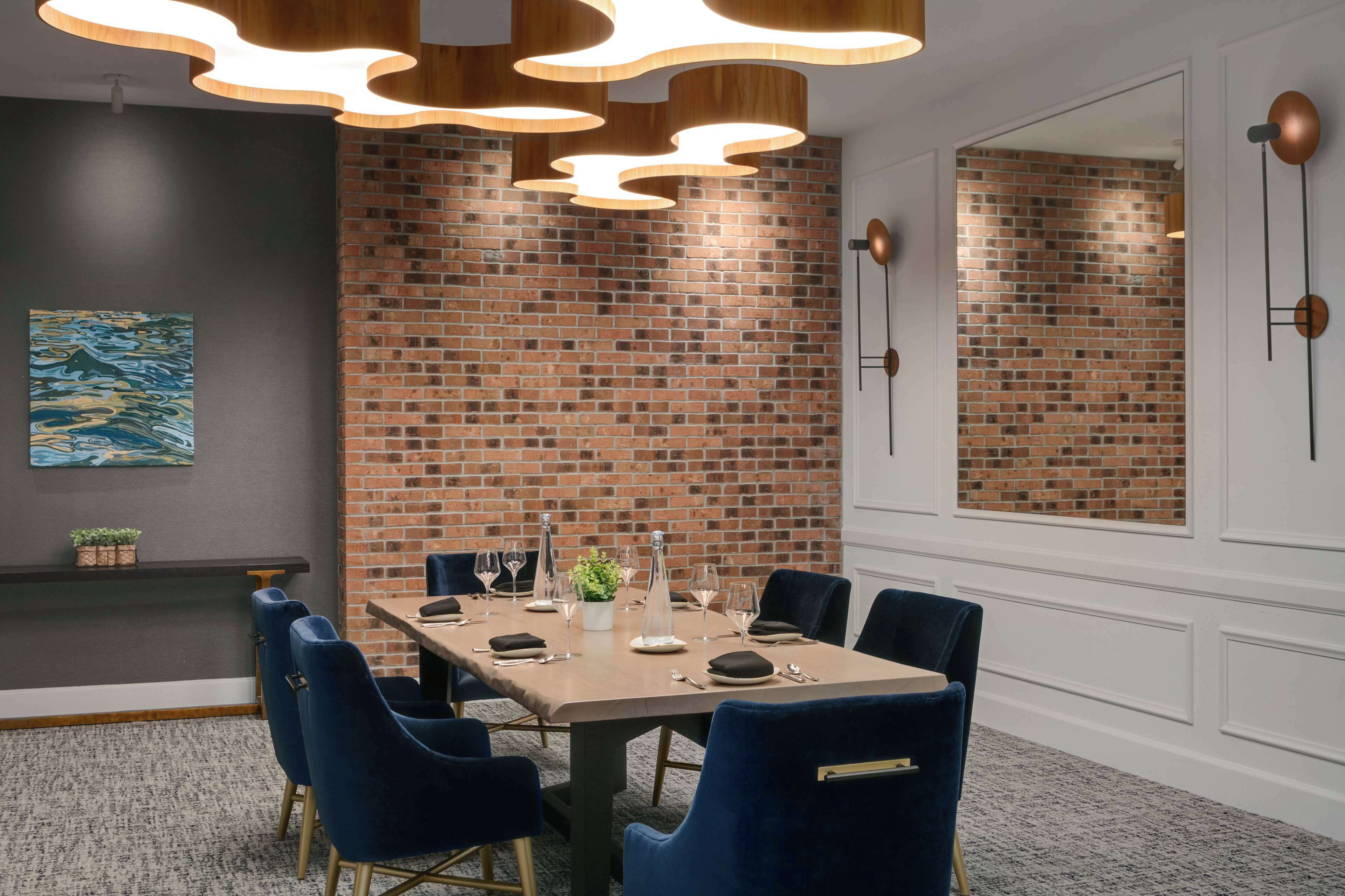 A conference room with a brick wall, a long table set for dining, blue chairs, and modern pendant lighting.