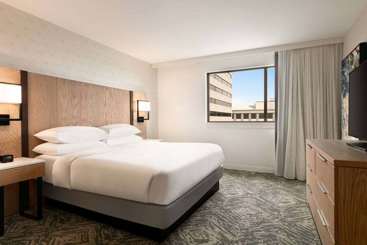 A hotel room with a large bed, a wooden headboard, a desk with a lamp, a dresser, and a window with a view of buildings.