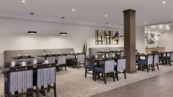 A modern hotel dining area with tables and chairs, a reception desk, and artwork on the walls.