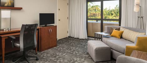 A hotel room with a desk, chair, television, sofa, and a view of greenery through the window.