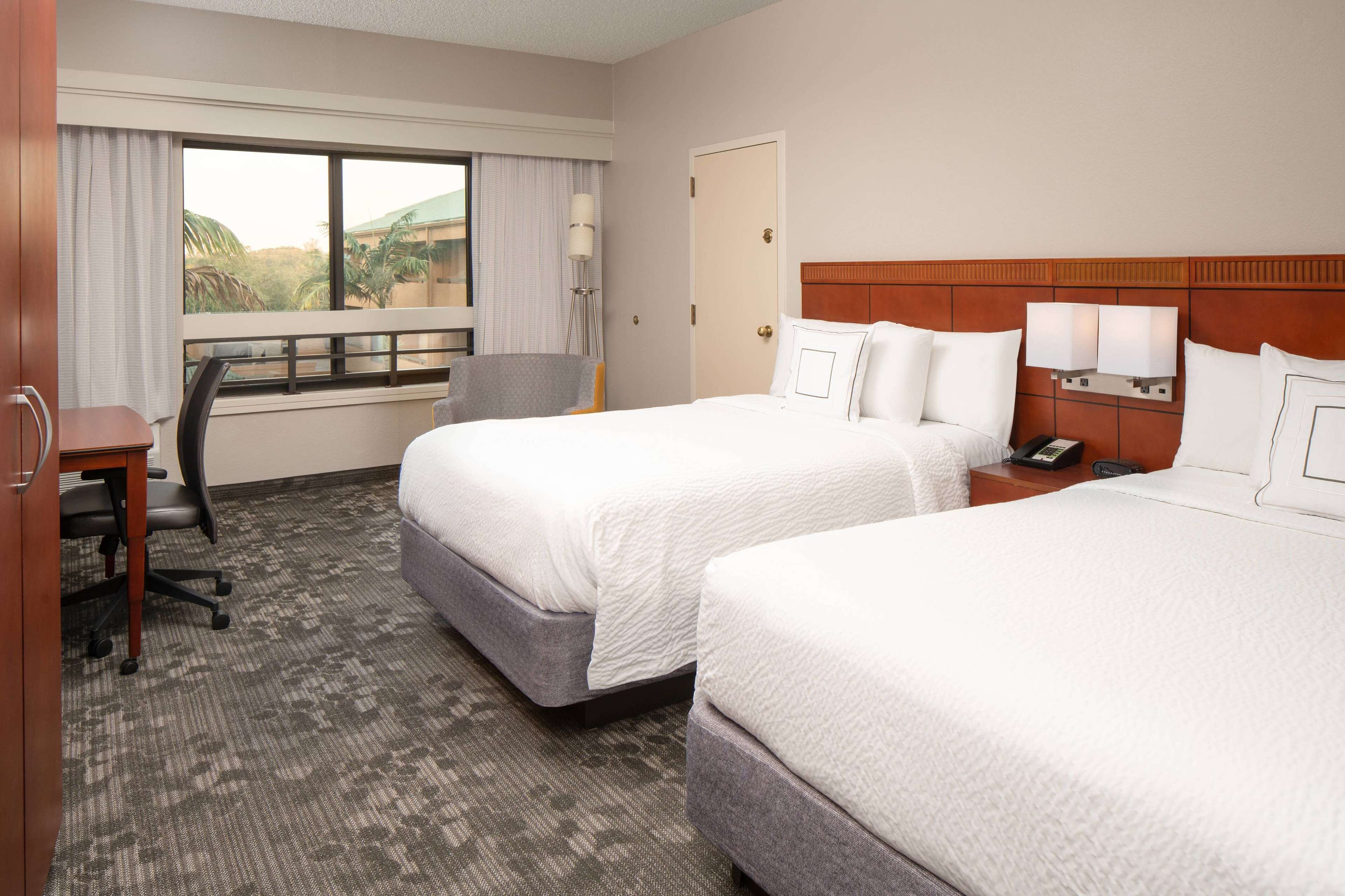 A hotel room with two beds, a desk, a chair, a window with curtains, and a view of palm trees.