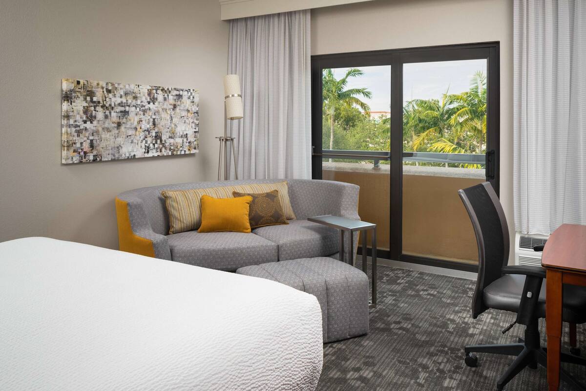 A hotel room with a bed, a grey sofa with yellow cushions, a small table, a chair, and a large window with a view of palm trees.