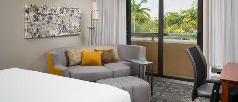 Ein Hotelzimmer mit einem Bett, einem grauen Sofa mit gelben Kissen, einem kleinen Tisch, einem Stuhl und einem großen Fenster mit Blick auf Palmen.