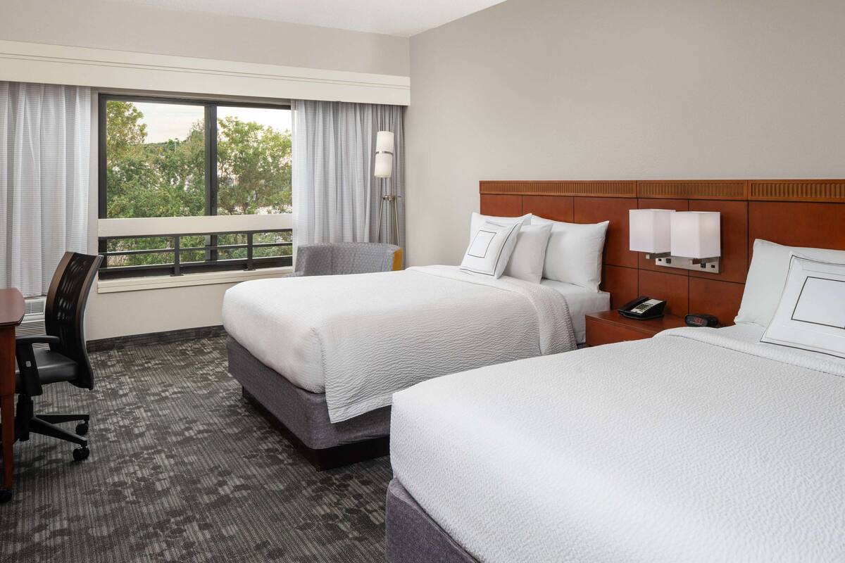 A hotel room with two beds, a desk, a chair, a nightstand, and a window with curtains.