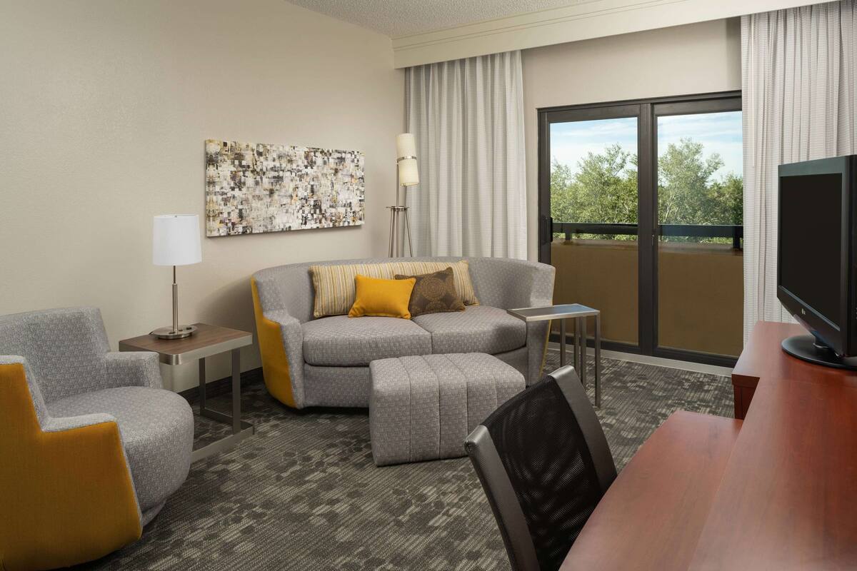A hotel room with a grey sofa, a small table, a TV, and a window with a view of trees.