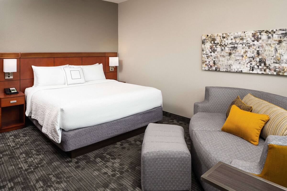 A hotel room with a large bed, a grey sofa, a small ottoman, and a wall-mounted artwork.