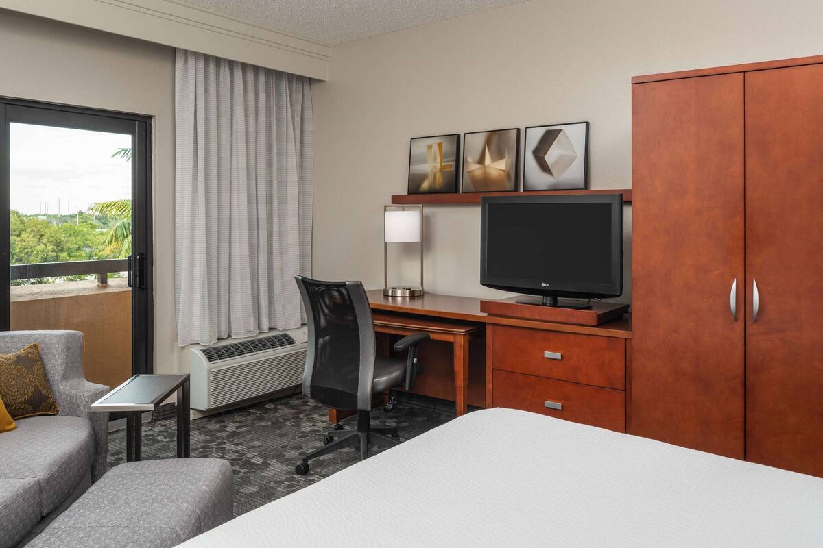 A hotel room with a bed, a desk with a TV, a chair, a small table, a heater, and a window with curtains.