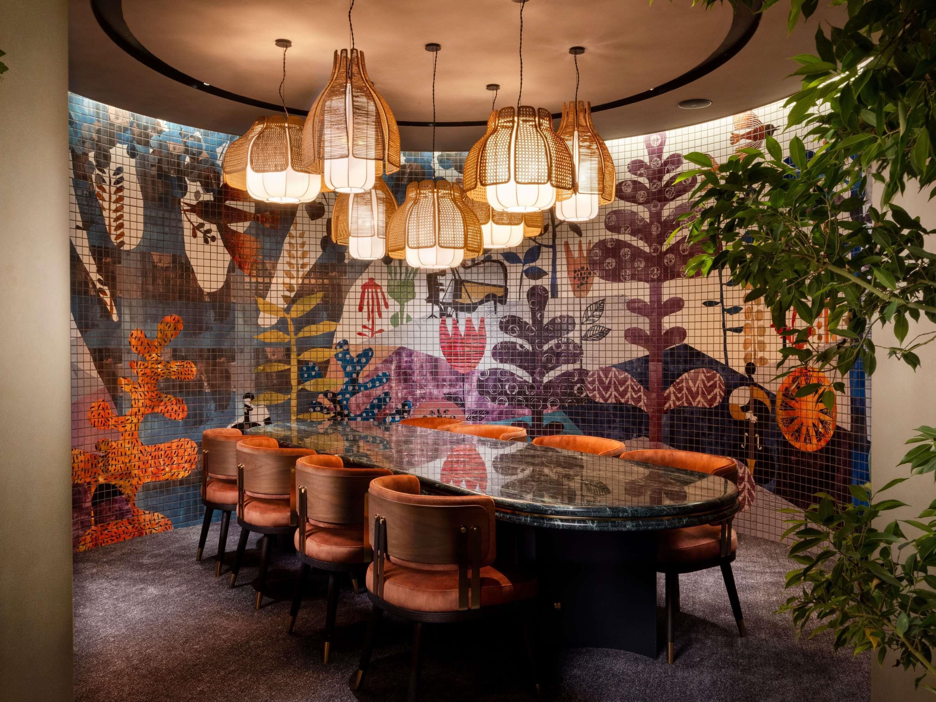 A dining area with a round table, chairs, hanging lights, and a colorful mosaic wall.