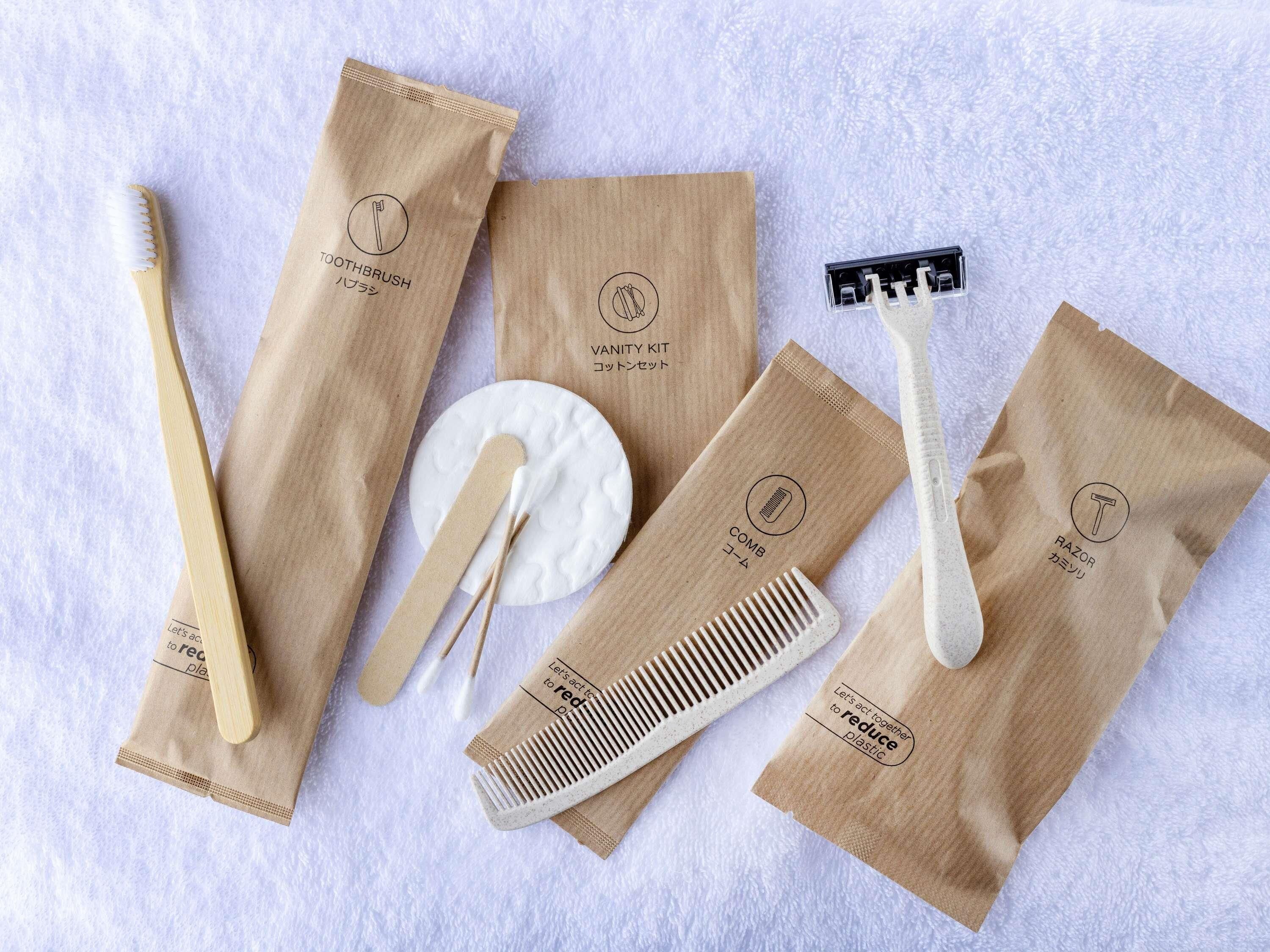 A set of personal care items including a toothbrush, razor, comb, and cotton swab, all packaged in brown paper bags with eco-friendly messages.