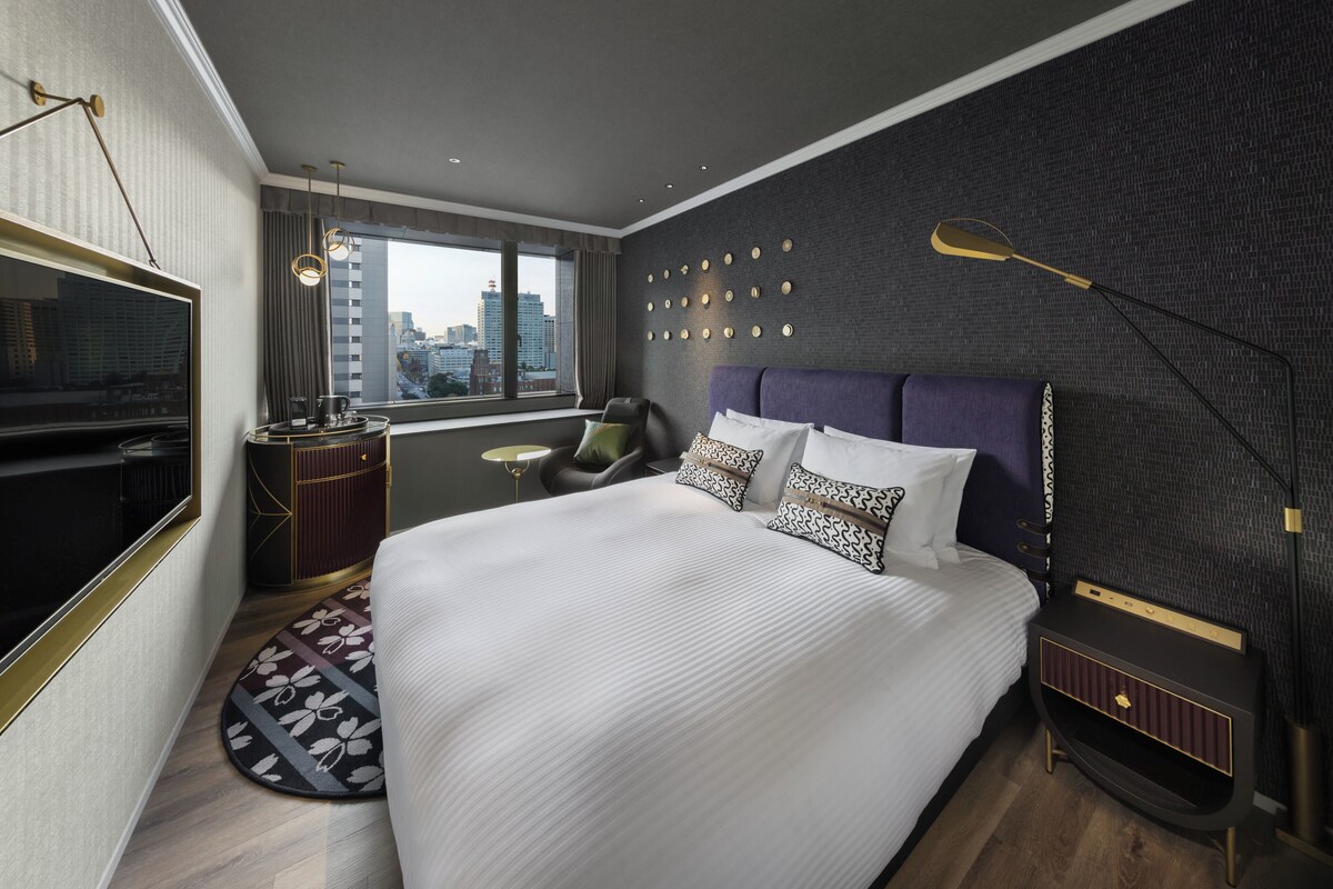 A modern hotel room with a large bed, a TV, a desk, and a city view.