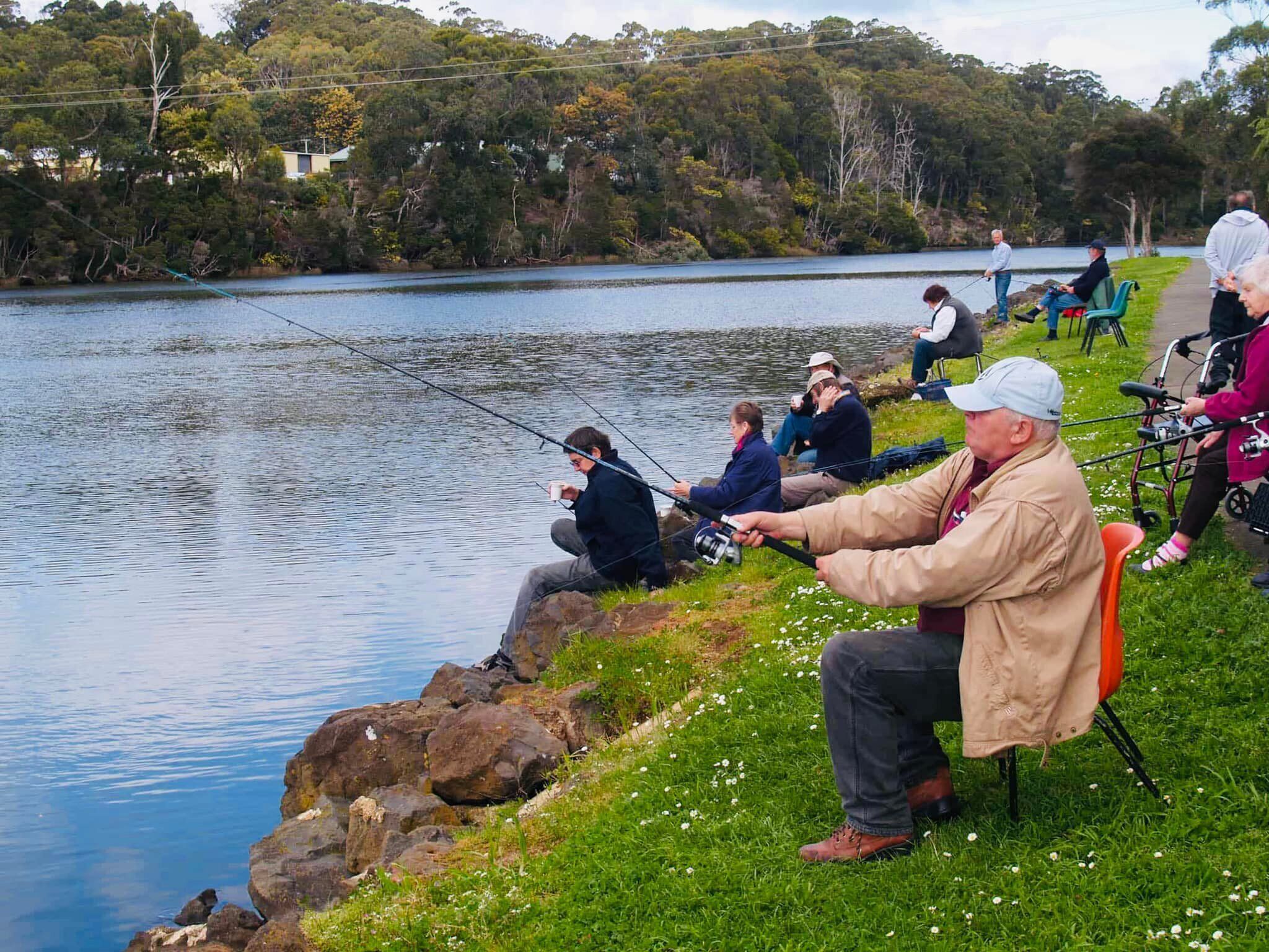 Zona de pesca junto al lago, con personas pescando, una orilla cubierta de césped y un paisaje de árboles y colinas.