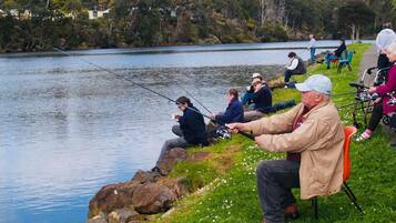 Zona de pesca junto al lago, con personas pescando, una orilla cubierta de césped y un paisaje de árboles y colinas.