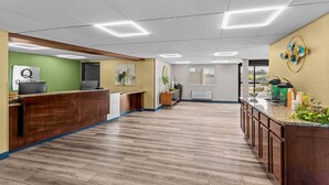 A reception area with a wooden reception desk, a green wall with a 'Quality' sign, a large window, and a kitchen counter with various items.