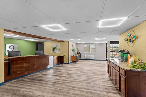 A reception area with a wooden reception desk, a green wall with a 'Quality' sign, a large window, and a kitchen counter with various items.