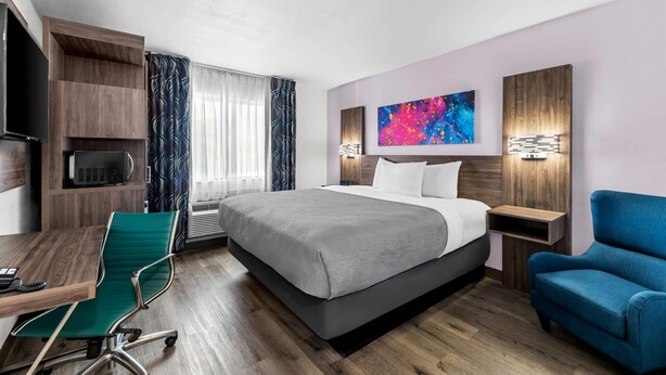 A modern hotel room with a large bed, a desk with a chair, a TV, and a blue armchair.