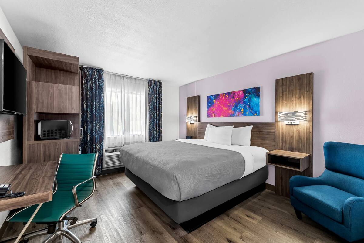 A modern hotel room with a large bed, a desk with a chair, a TV, and a blue armchair.
