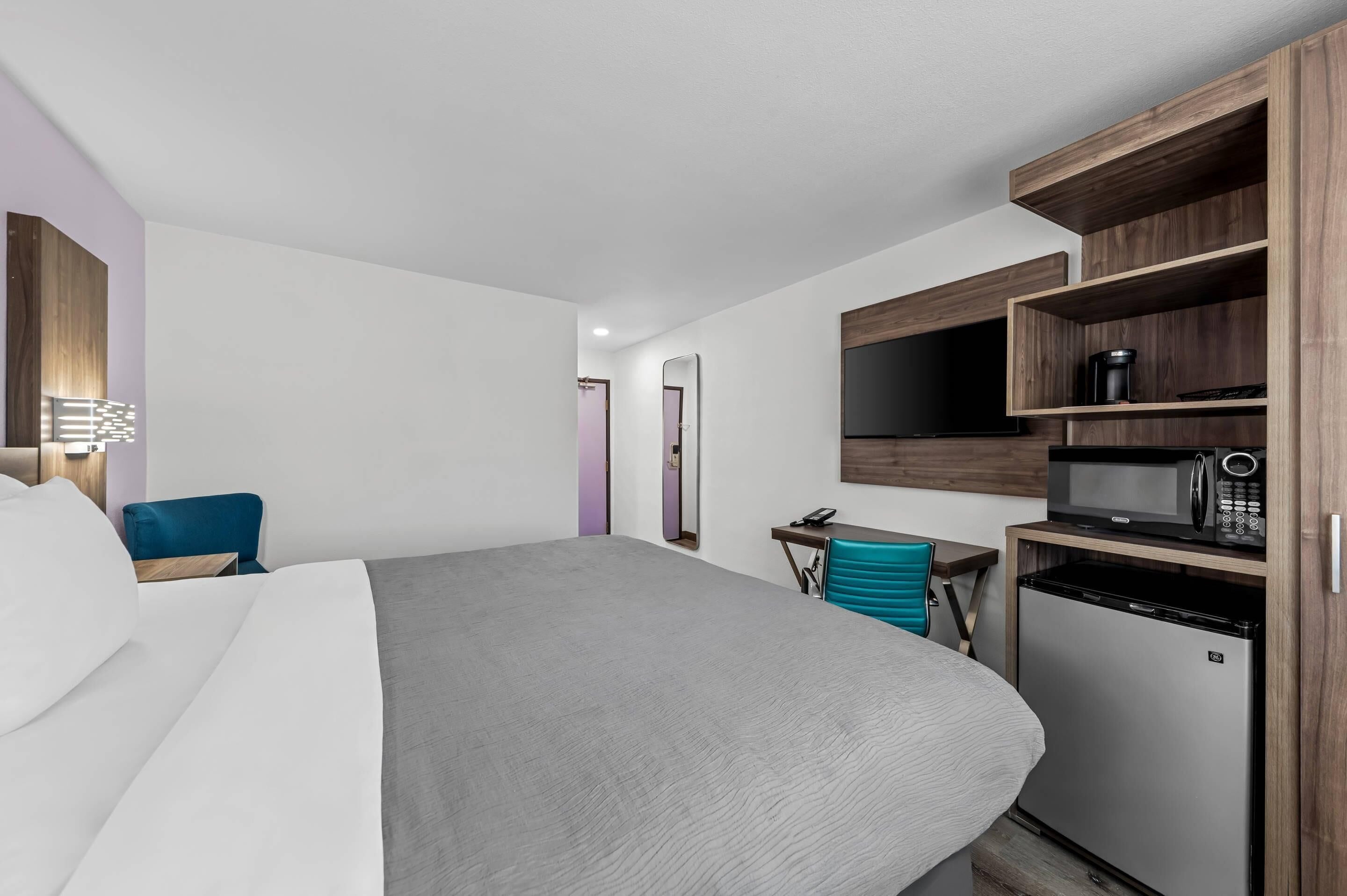 A modern hotel room with a large bed, a desk with a chair, a microwave, a refrigerator, and a TV mounted on the wall.
