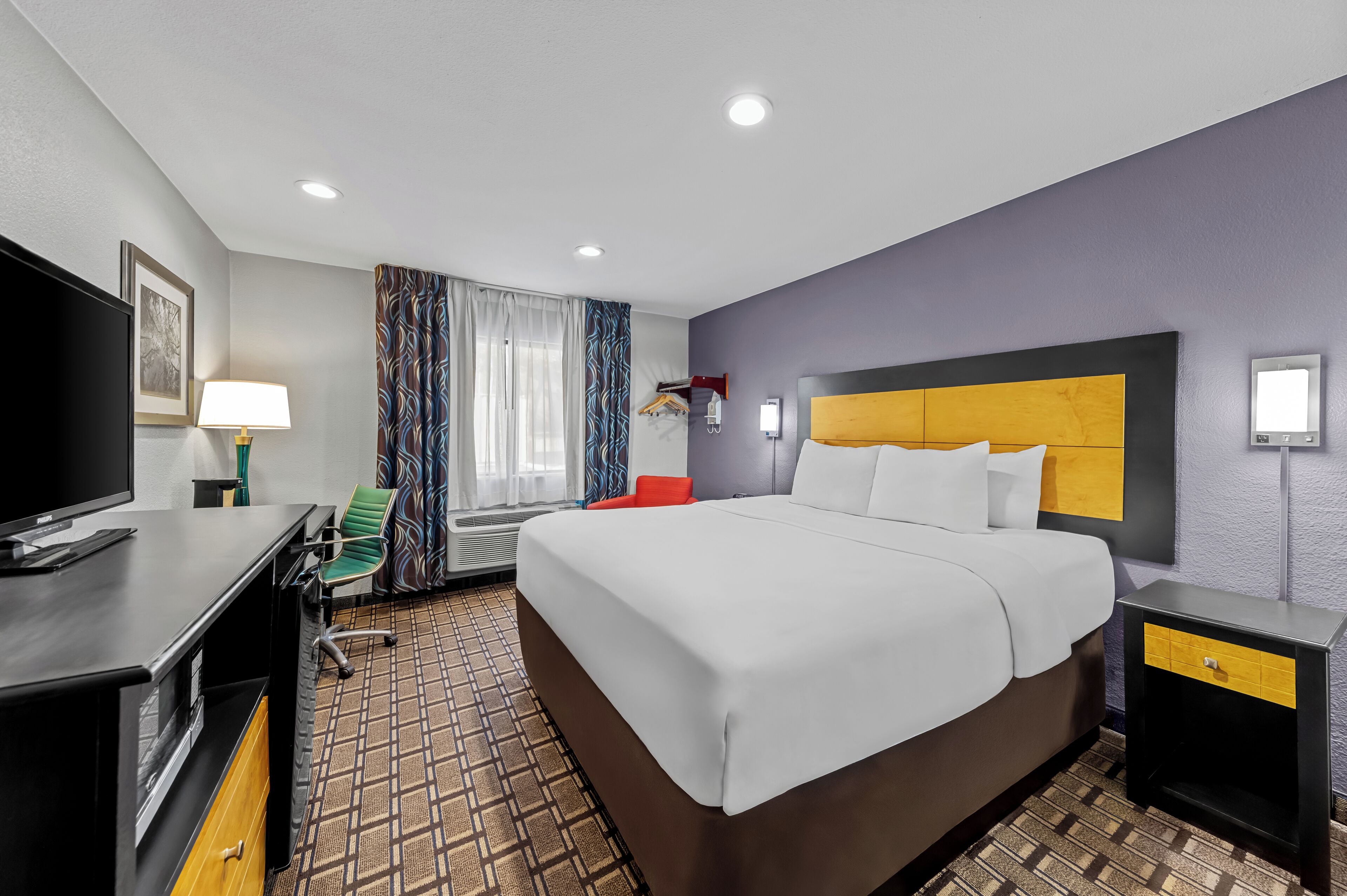 A hotel room with a large bed, a desk with a TV, a chair, a wardrobe, and a bedside table.