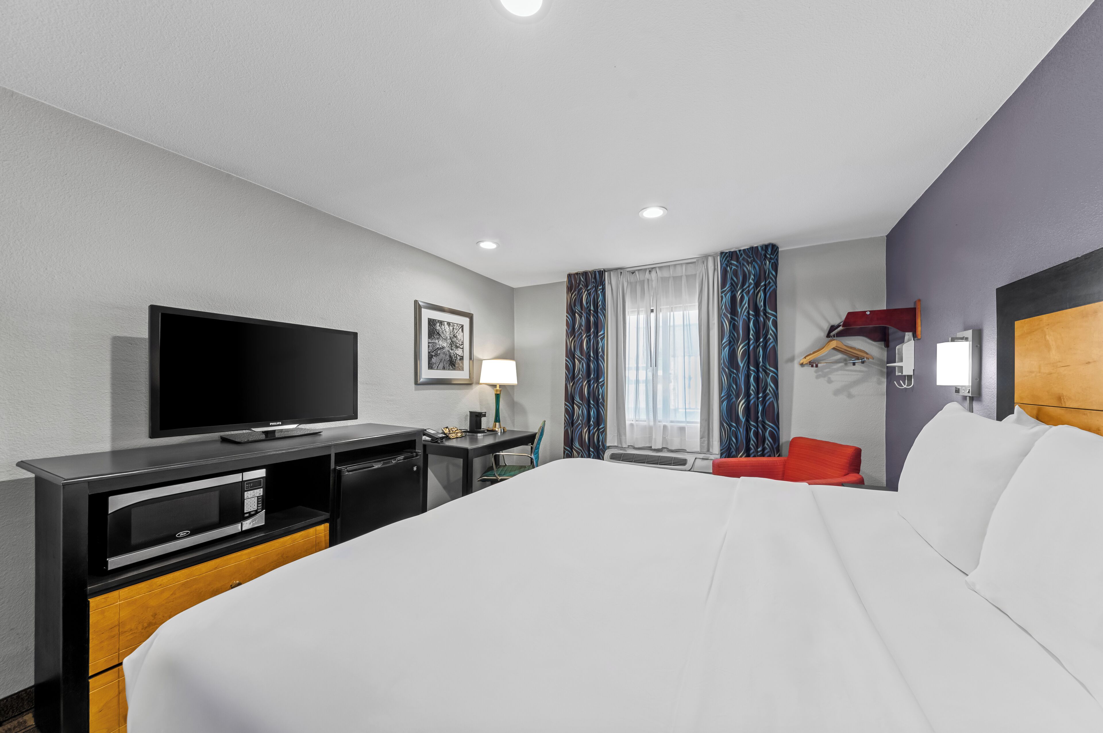 A hotel room with a large bed, a flat-screen TV, a desk with a lamp, a chair, and a window with curtains.