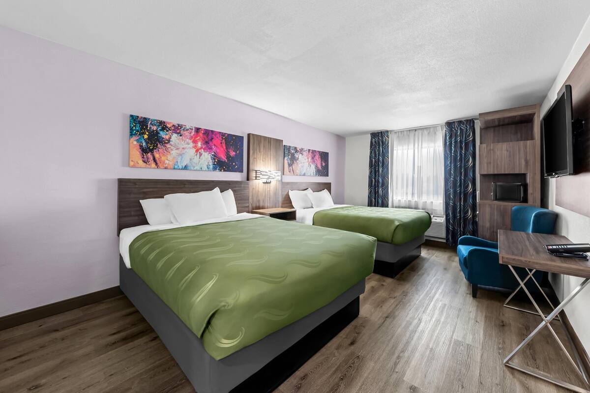 A hotel room with two beds, a desk, a chair, a TV, and artwork on the walls.