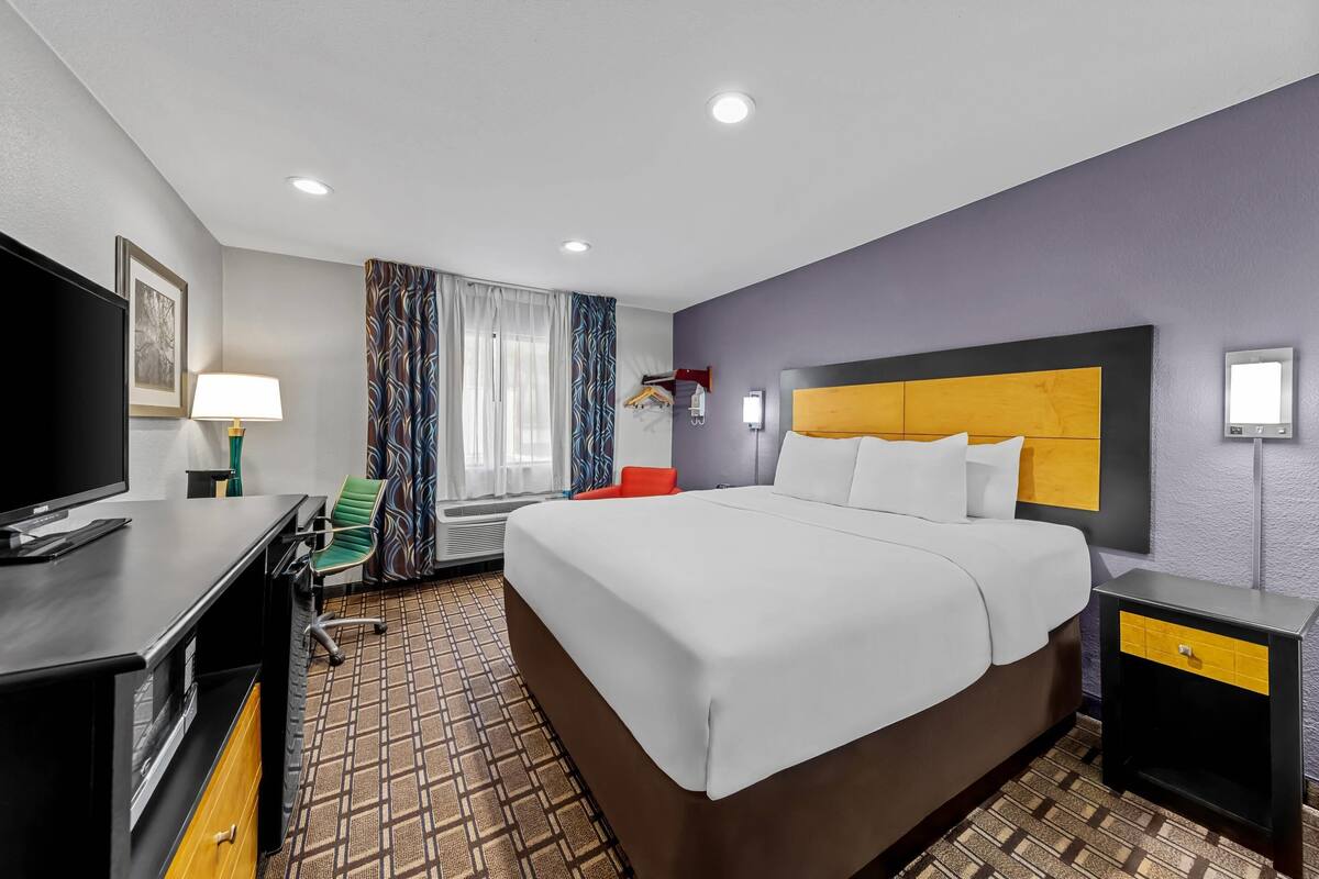 A hotel room with a large bed, a desk with a TV, a chair, and a bedside table with a lamp.