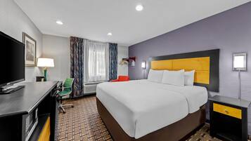 A hotel room with a large bed, a desk with a TV, a chair, and a bedside table with a lamp.