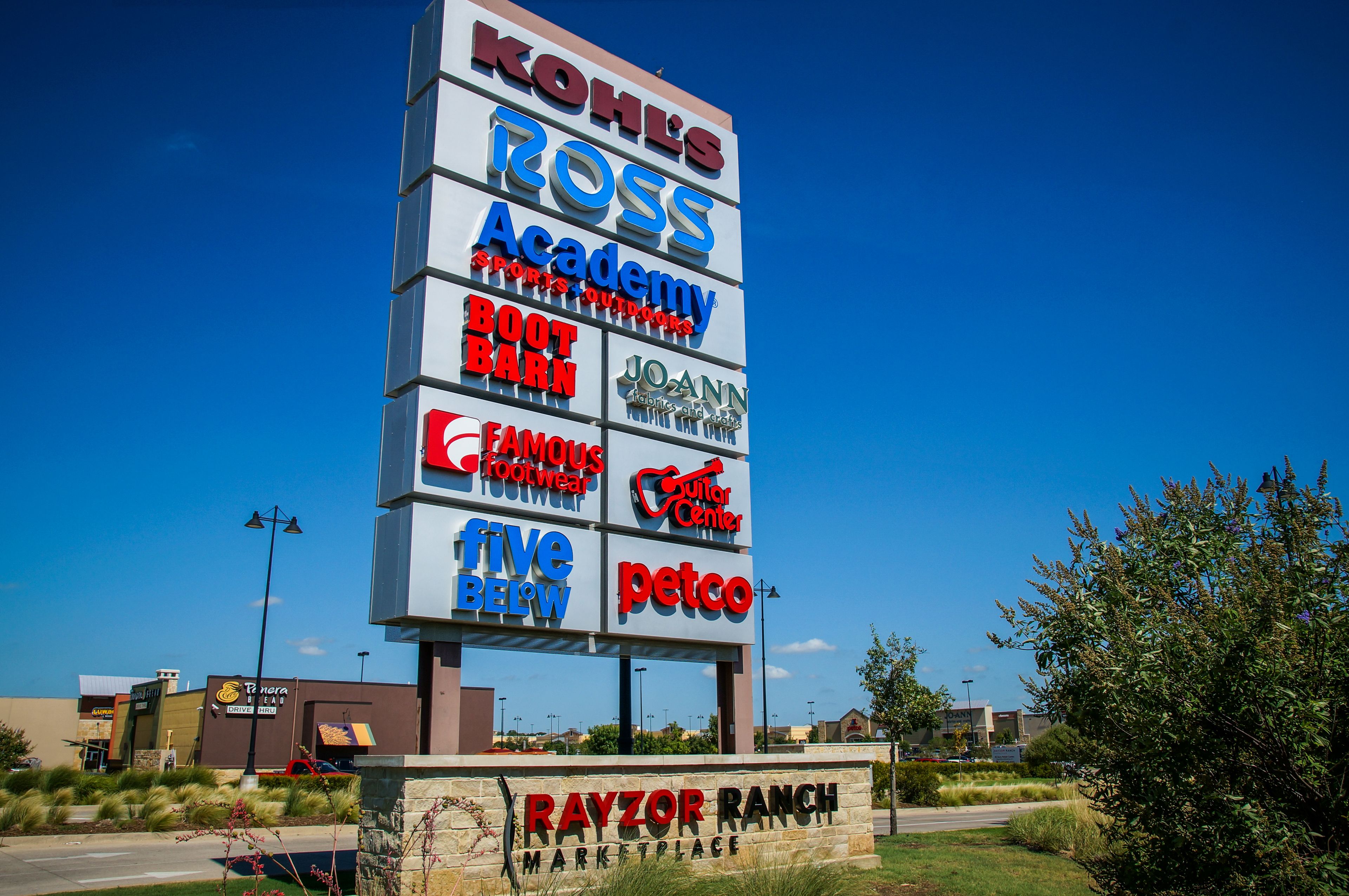 Un cartel comercial con varios nombres de marcas y logotipos, incluyendo Kohl's, Ross y Petco, sobre un cielo azul despejado.