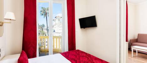 A hotel room with a bed, a TV on the wall, and a balcony with a view of palm trees.