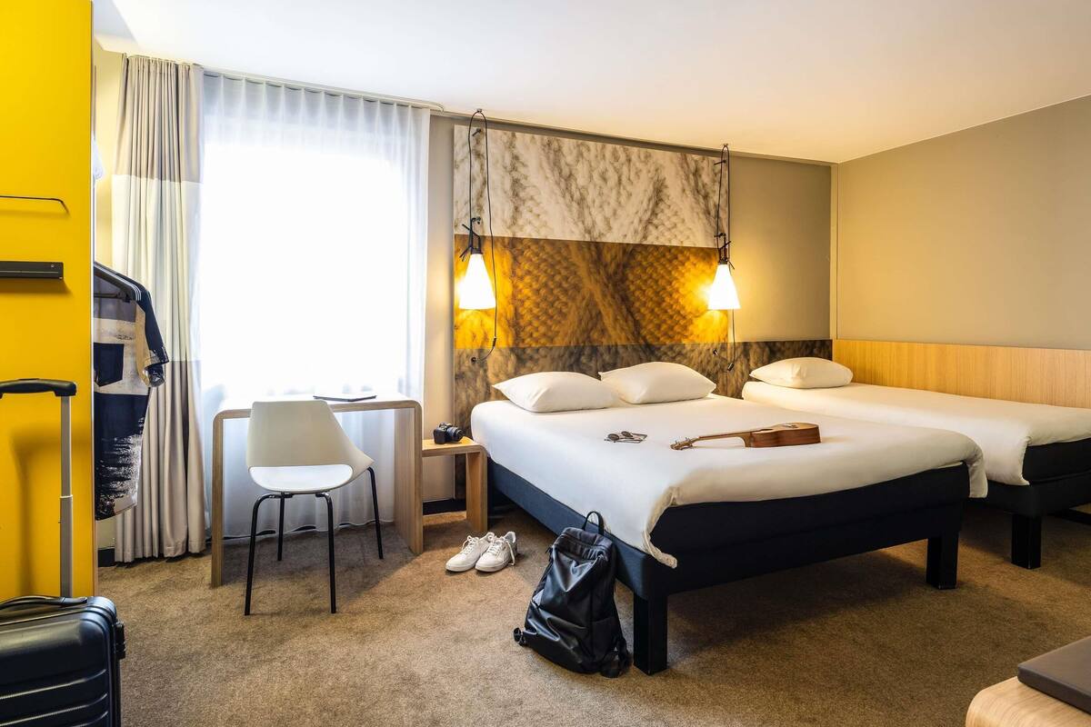 Quarto de hotel com duas camas, uma escrivaninha, uma cadeira e uma mala no chão.