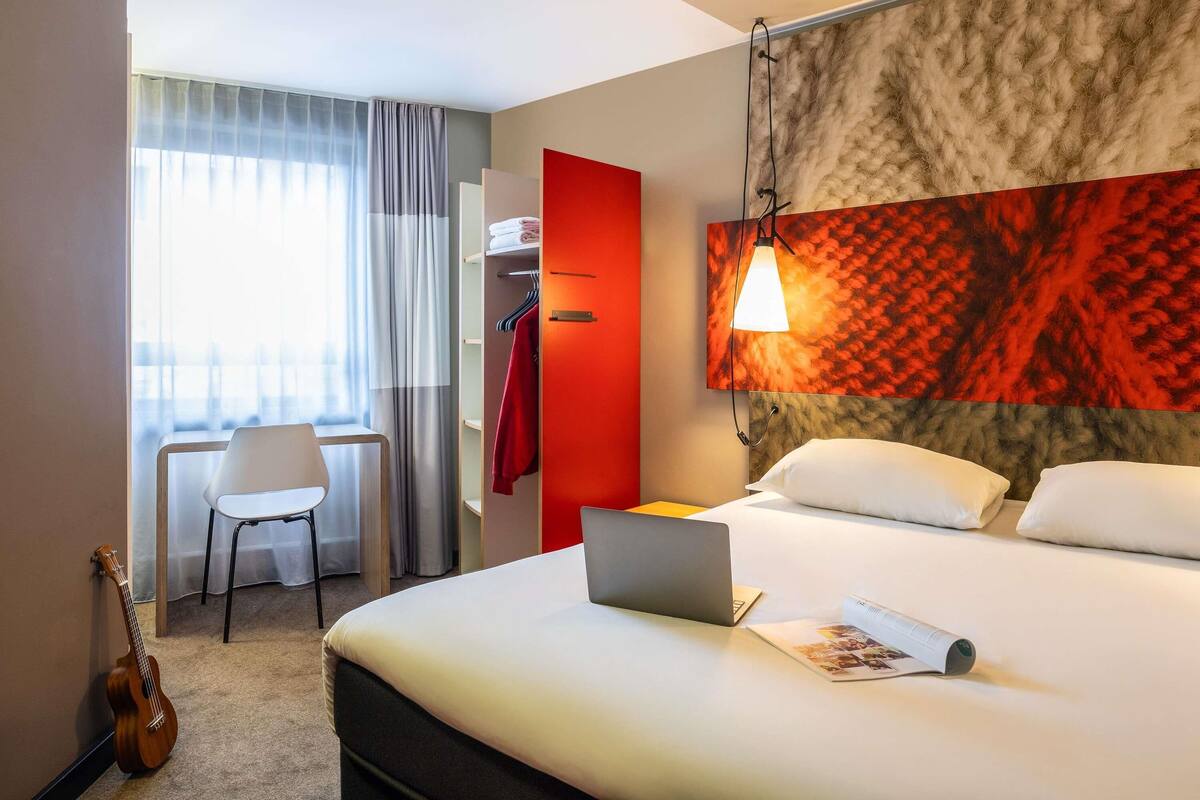 Quarto de hotel com cama, um laptop, uma guitarra, escrivaninha com cadeira e uma porta vermelha.