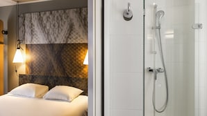 Habitación de hotel con cama, dos almohadas, cabecera con diseño texturizado, ducha con alcachofa de mano e iluminación empotrada en la pared.